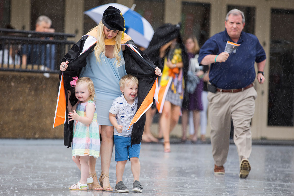 Mother, Dr. Lauren Hannah, protects her children from the rain with her gown while leaving the College of Social Work Commencement inside Thompson-Boling Arena
